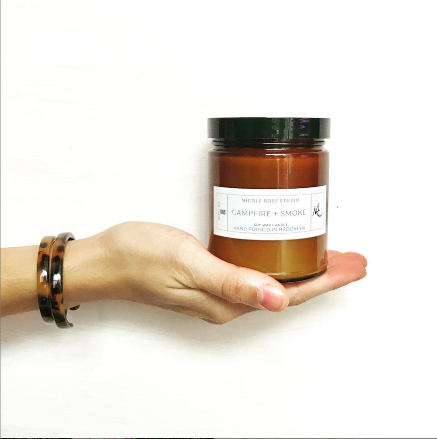 Hand holding campfire smoke soy candle in amber jar, rustic cozy scent.
