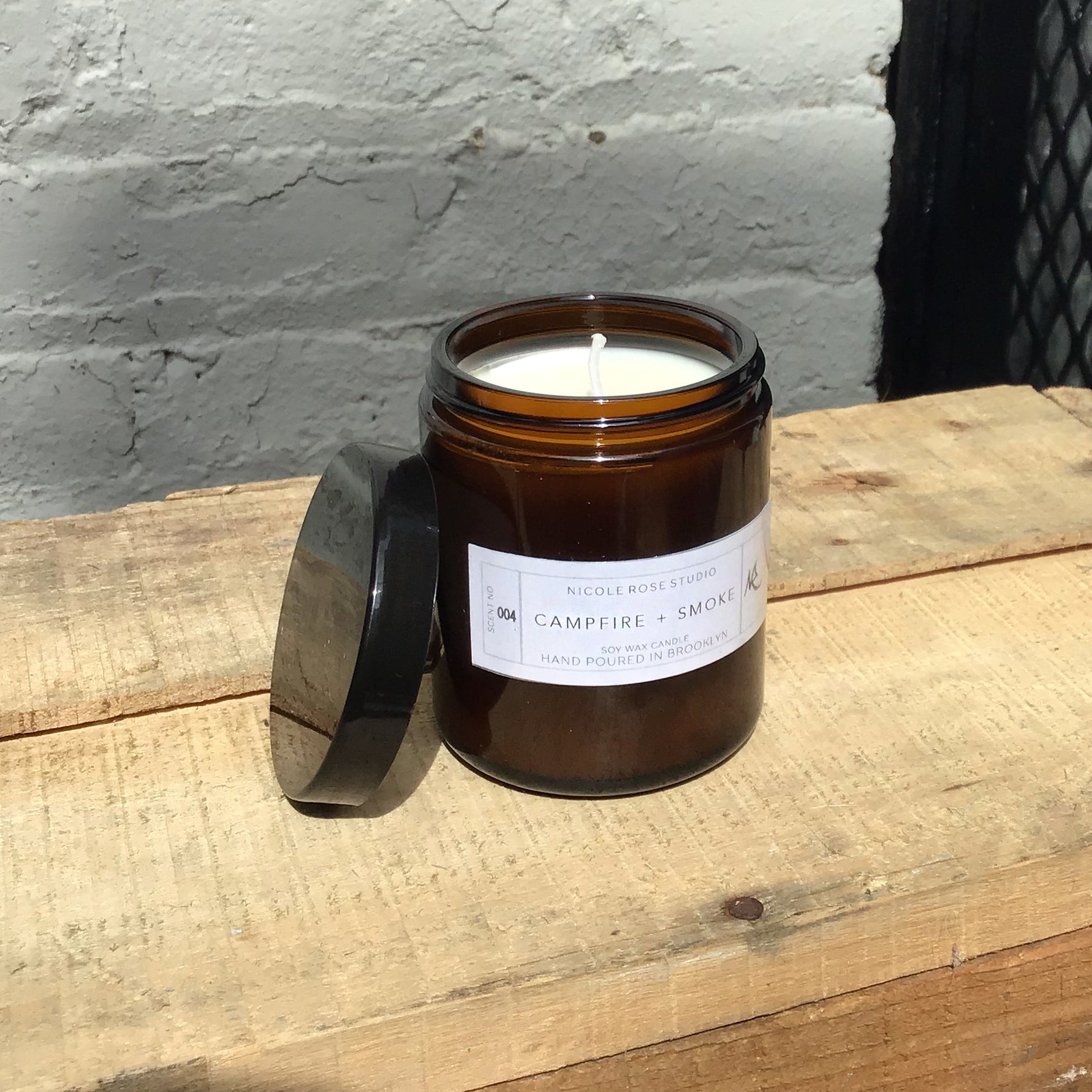 Campfire smoke soy candle in amber jar with lid, cozy rustic aroma.
