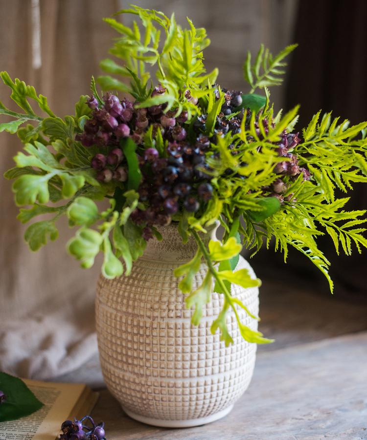 Textured ceramic vase with green foliage and purple berries for natural home decor.
